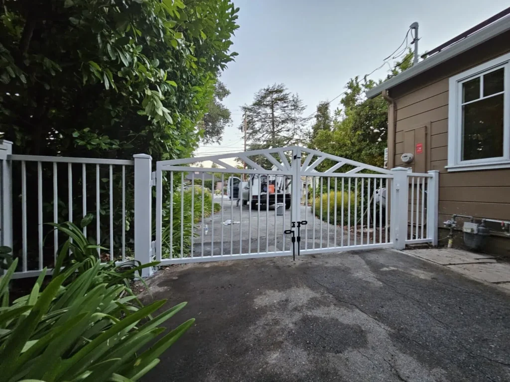 White metal double swing driveway gate installed at a residential property with trees, a paved driveway, and a house nearby.