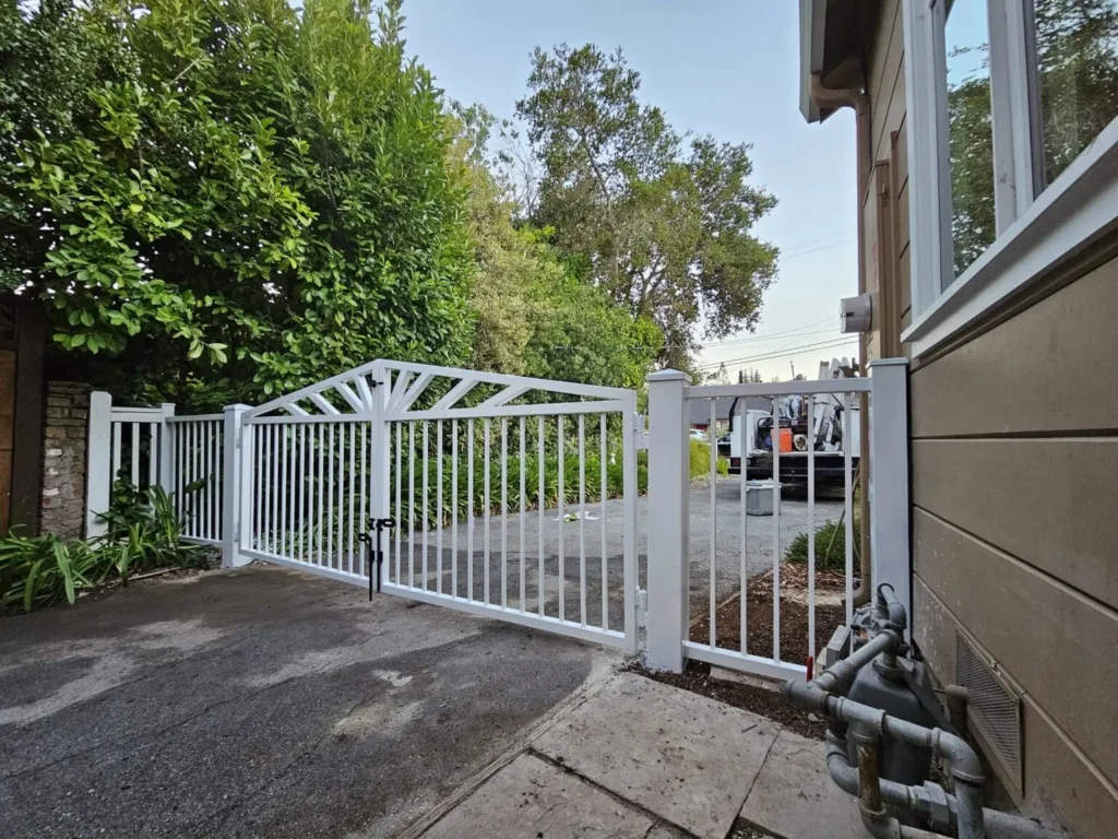 White metal swing driveway gate with vertical bars installed beside a house and landscaped front yard.
