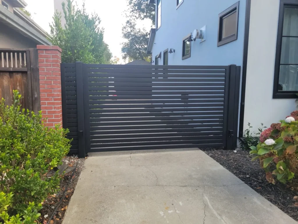 Modern black horizontal slat driveway gate installed at a contemporary home with a concrete driveway and surrounding greenery.