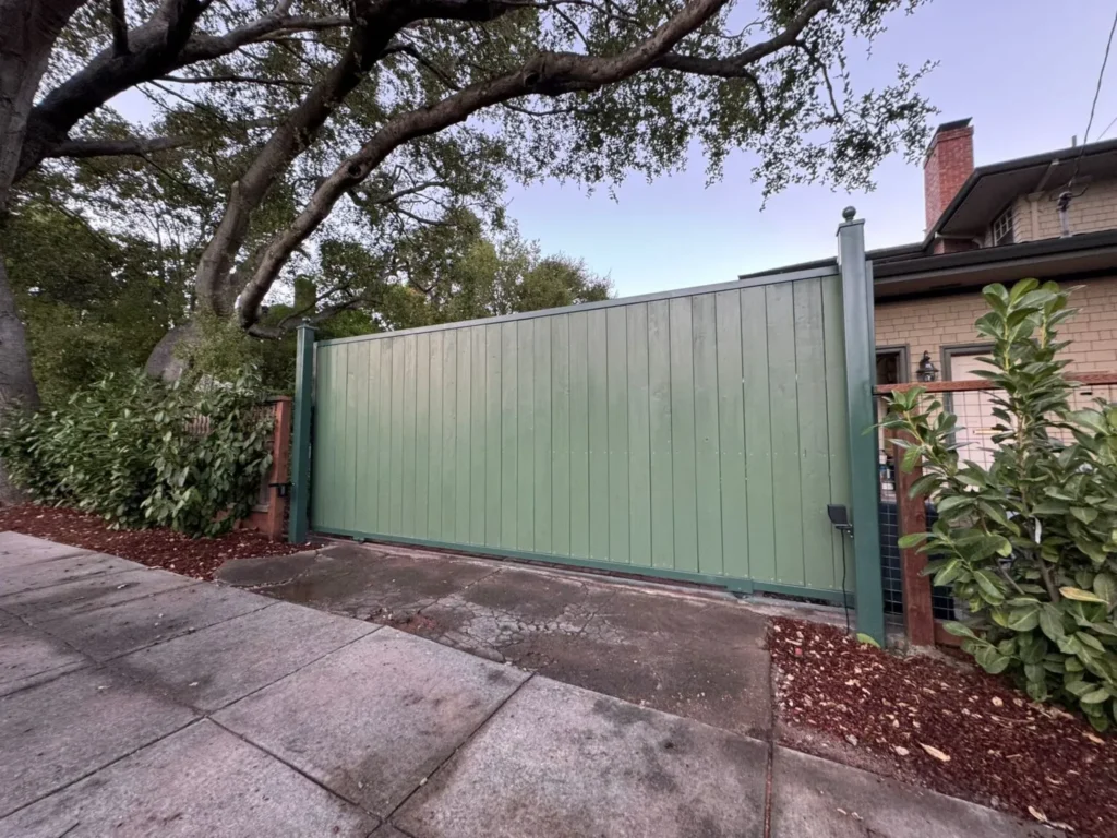 Green metal sliding driveway gate installed beneath a large tree beside a residential property.