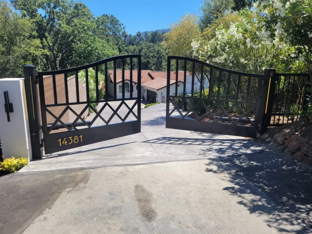 Decorative black metal driveway gate with wood-look panels installed at a sloped residential entrance surrounded by trees.